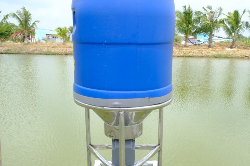 Cận cảnh thùng chứa và cổ phễu inox của máy cho tôm ăn FarmX A30 dựng bên bờ ao, nền là mặt nước và hàng dừa