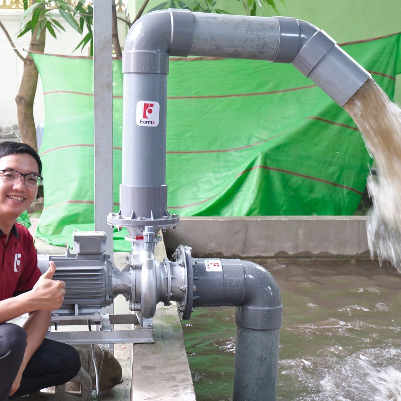 Người nuôi tôm ngồi cạnh máy bơm nước FarmX F1 MG168 liền mô tơ đang chạy, nước xả ra ao, xưởng FarmX Đầm Dơi Cà Mau
