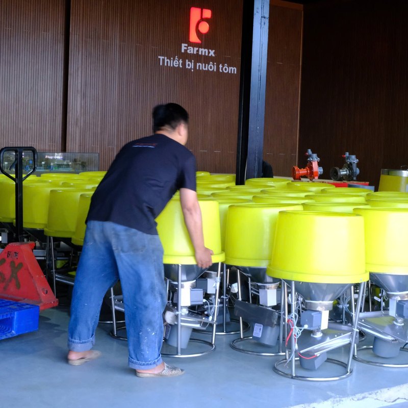 Máy cho tôm ăn FarmX XP50 trong kho xưởng Đầm Dơi, Cà Mau, nhìn từ góc khác thấy khung inox và phễu vàng