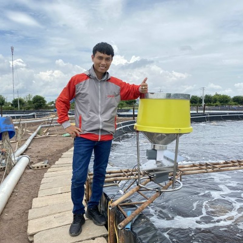 Máy cho tôm ăn FarmX XP50 đặt mé bờ ao tôm nhìn ngang, thấy chân đế, phễu vàng và mô tơ gắn dưới đáy